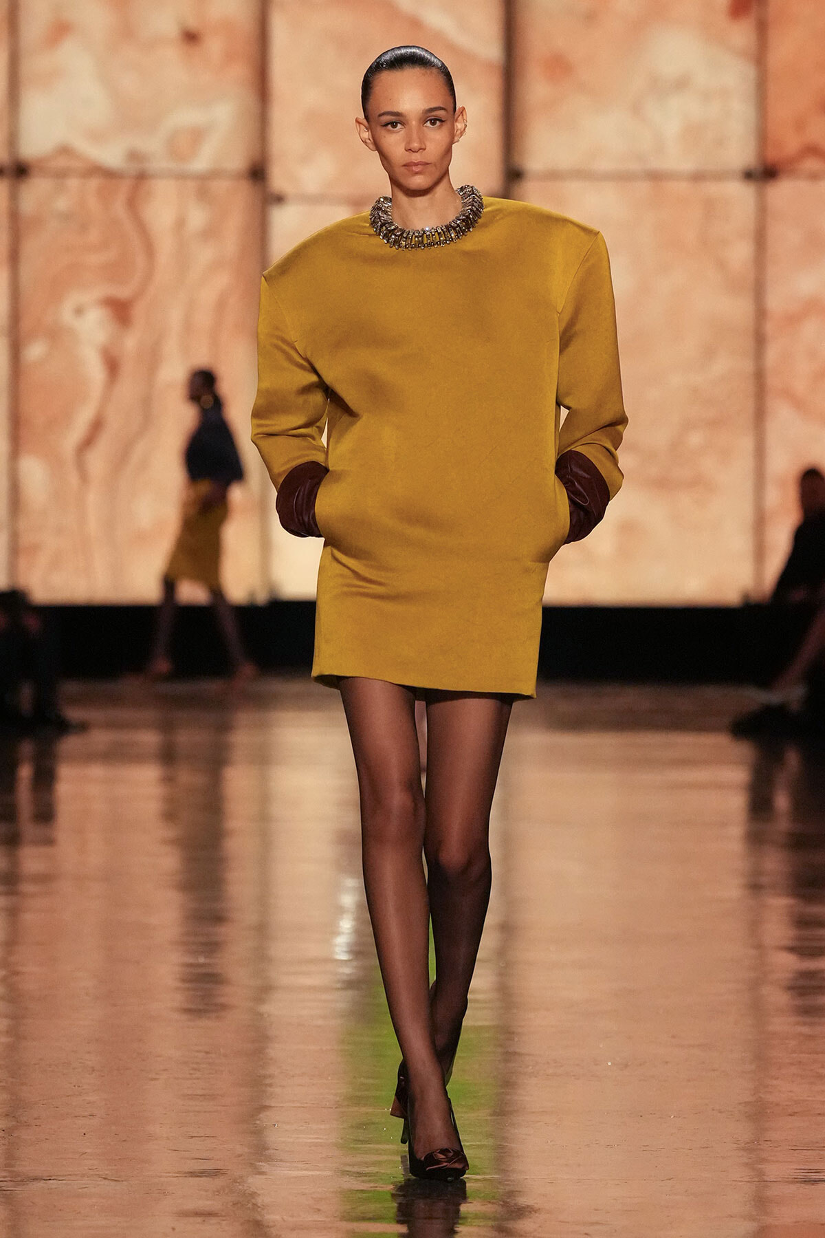 Model walking a fashion runway in a mustard-yellow mini dress with brown cuffed sleeves, hands in pockets, paired with sheer black tights, black heels, and a chunky beaded collar necklace.