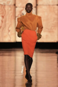 Model walking runway in mustard turtleneck top with oversized sleeves, paired with orange pencil skirt, black tights, and dark heeled shoes; gold statement earrings.