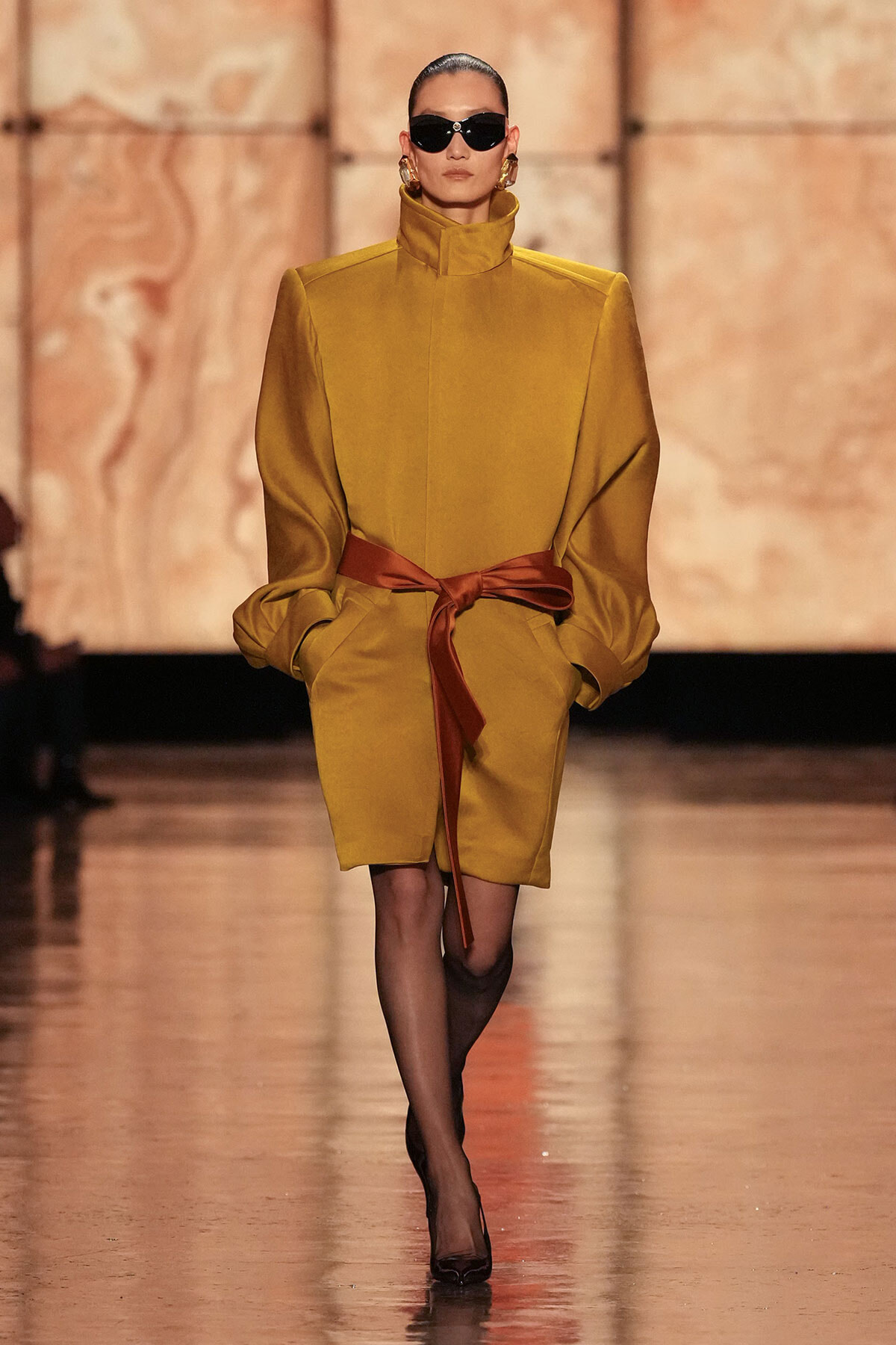 Runway model in a mustard-yellow coat with a high collar, cinched by a brown sash belt, wearing black sunglasses and heels.