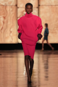 Model walks the runway in a hot-pink oversized turtleneck dress with dark gloves and gold earrings, against a beige backdrop.