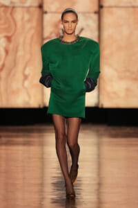 Model walking a fashion runway in a green mini dress with black gloves and a chunky metallic necklace.