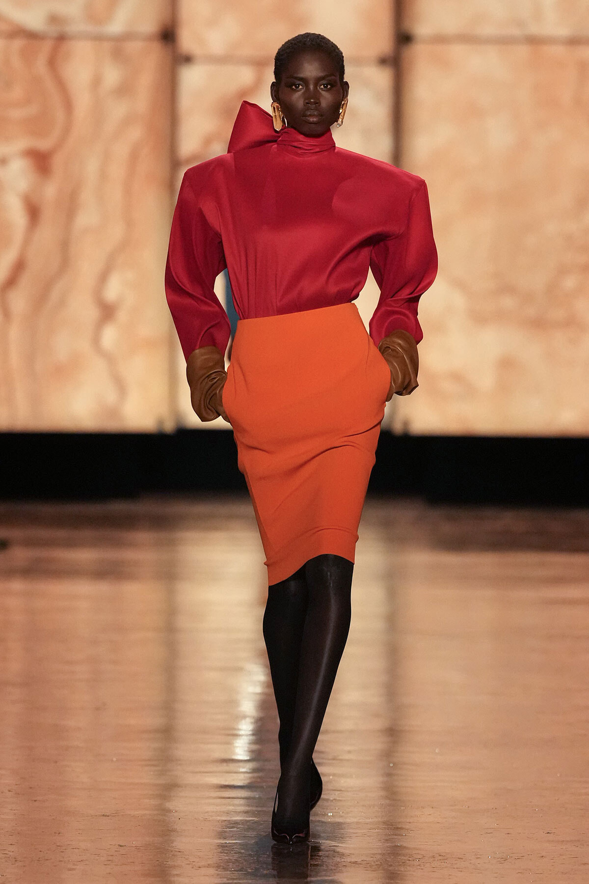 Model walks a runway in a red high-neck blouse with puff sleeves and an orange pencil skirt, black tights, brown gloves, and gold earrings.