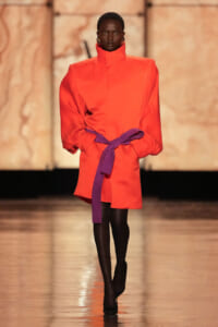 Runway model in a bright orange oversized coat with a purple belt tied at the waist, black tights and heels.