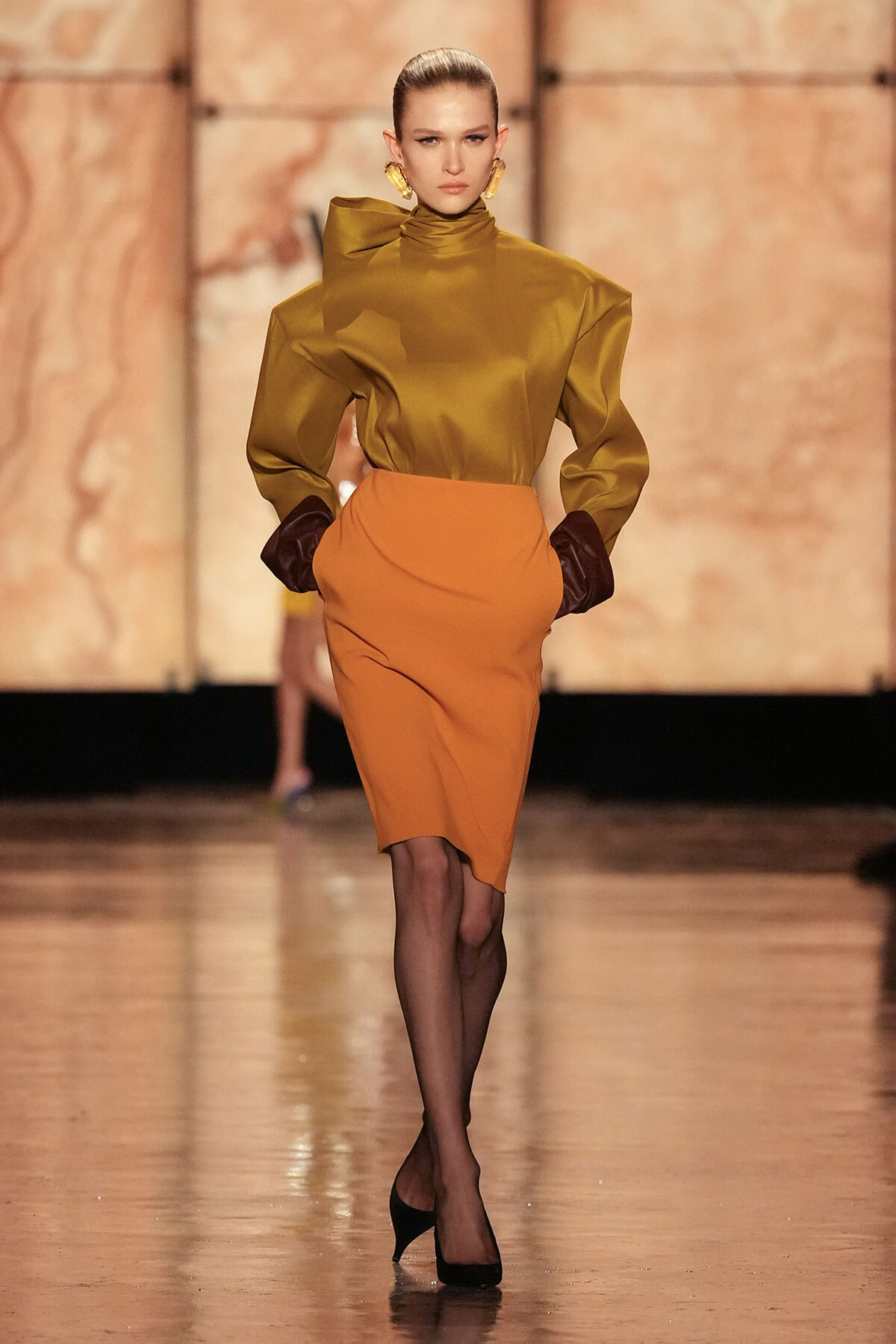 Model walking a fashion runway in a mustard high-neck blouse and orange pencil skirt, black gloves, and gold earrings.