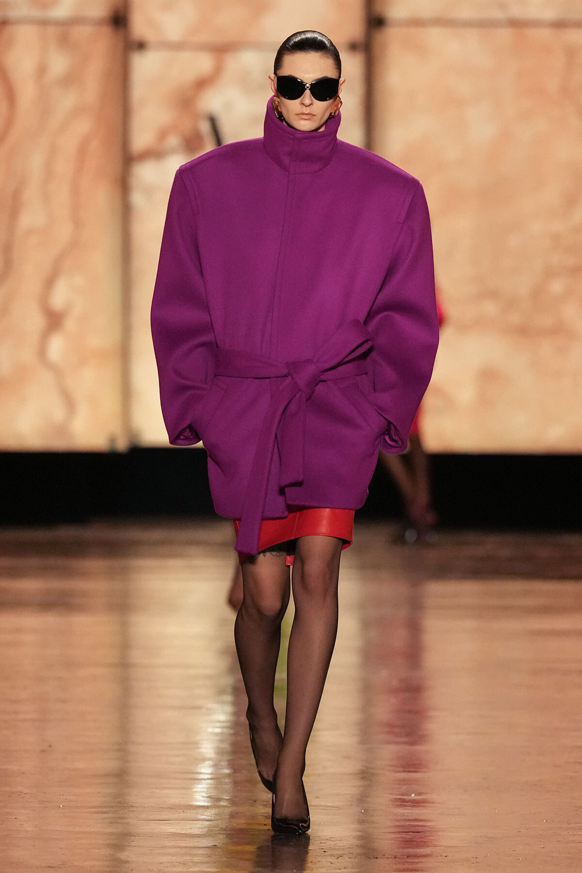 Model on a runway wearing an oversized purple coat with a high collar and tied belt, black sunglasses, and a red hem showing underneath.