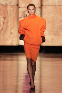 Model walks a runway in a bright orange monochrome outfit: a high‑neck top with puffed sleeves and a knee‑length skirt, black gloves at the wrists.