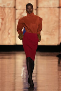 Model walks a fashion runway in a burnt orange turtleneck with sculpted shoulders and a burgundy pencil skirt, brown gloves, and black tights.
