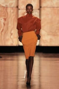 Model walking the runway in a rust-orange turtleneck blouse with structured shoulders and a high-waisted orange pencil skirt, paired with black gloves.