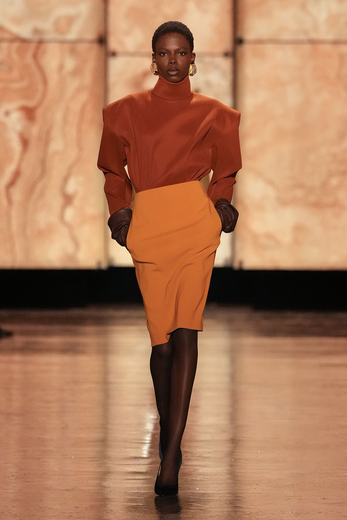 Model walking the runway in a rust-orange turtleneck blouse with structured shoulders and a high-waisted orange pencil skirt, paired with black gloves.