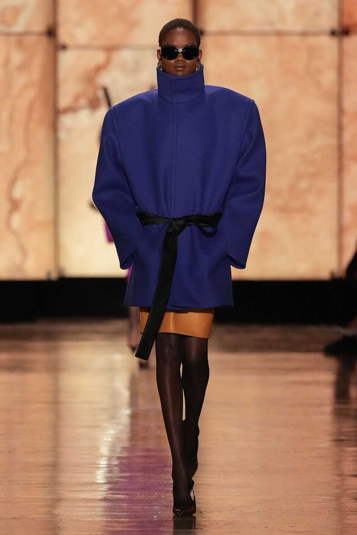 Model on a runway wearing an oversized royal blue coat with a high collar, cinched with a black belt, black sunglasses, and a tan mini skirt with black tights and heeled shoes.