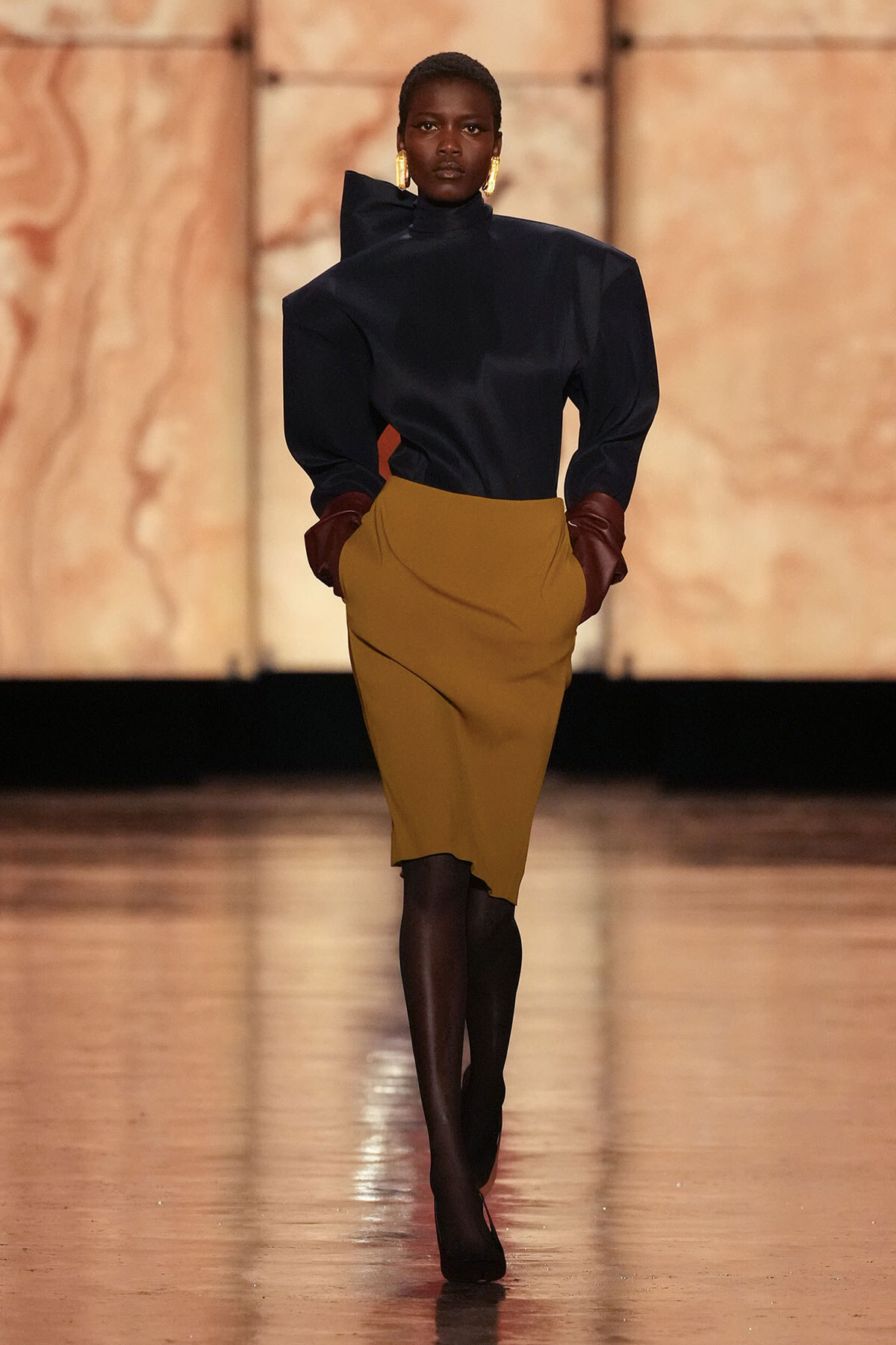 Fashion model on a runway wearing a navy high-neck top with structured shoulders, a mustard pencil skirt, maroon gloves, and gold earrings.