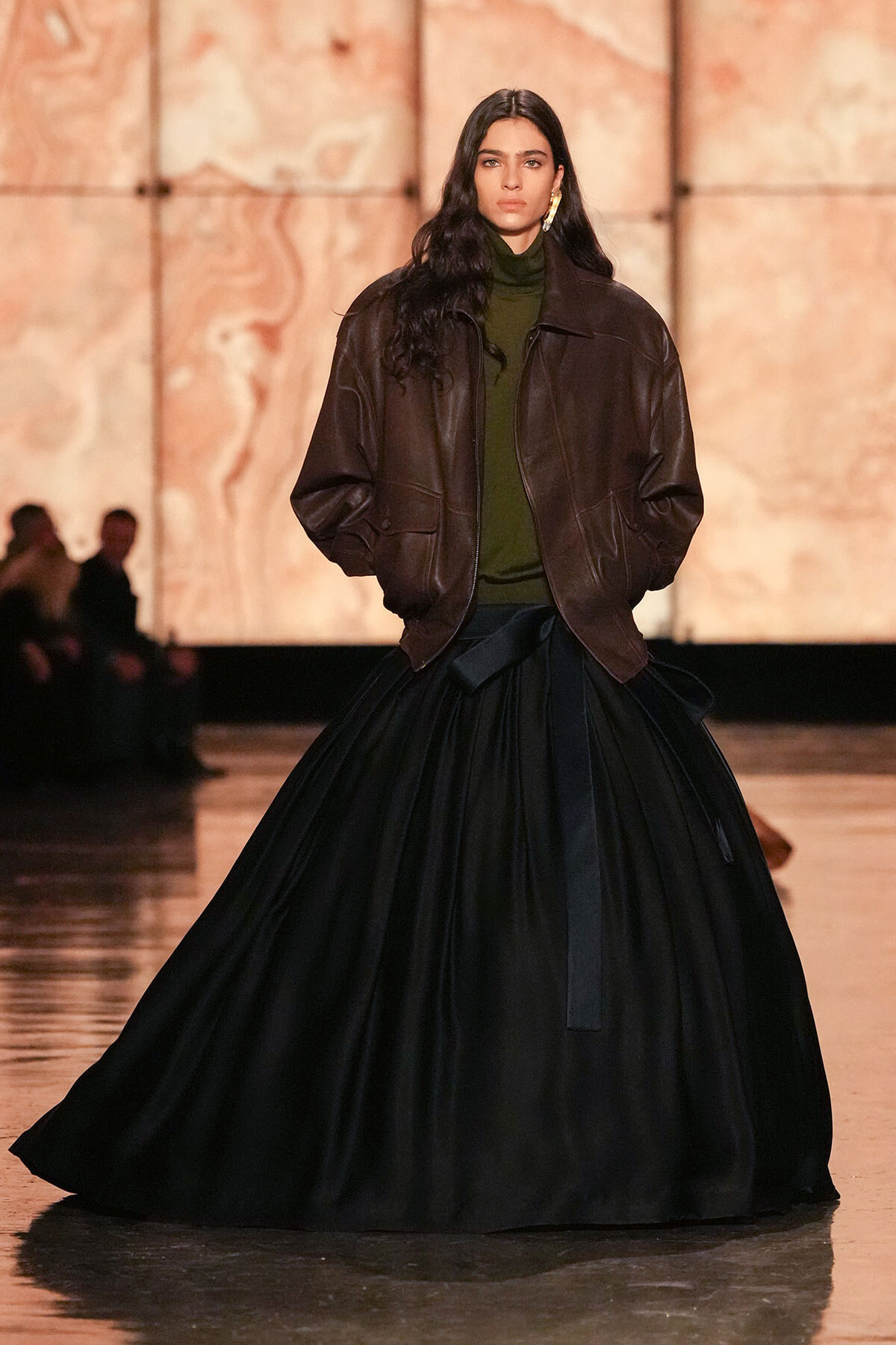 Model on runway wearing an oversized brown leather jacket, olive green turtleneck, and a voluminous black skirt.