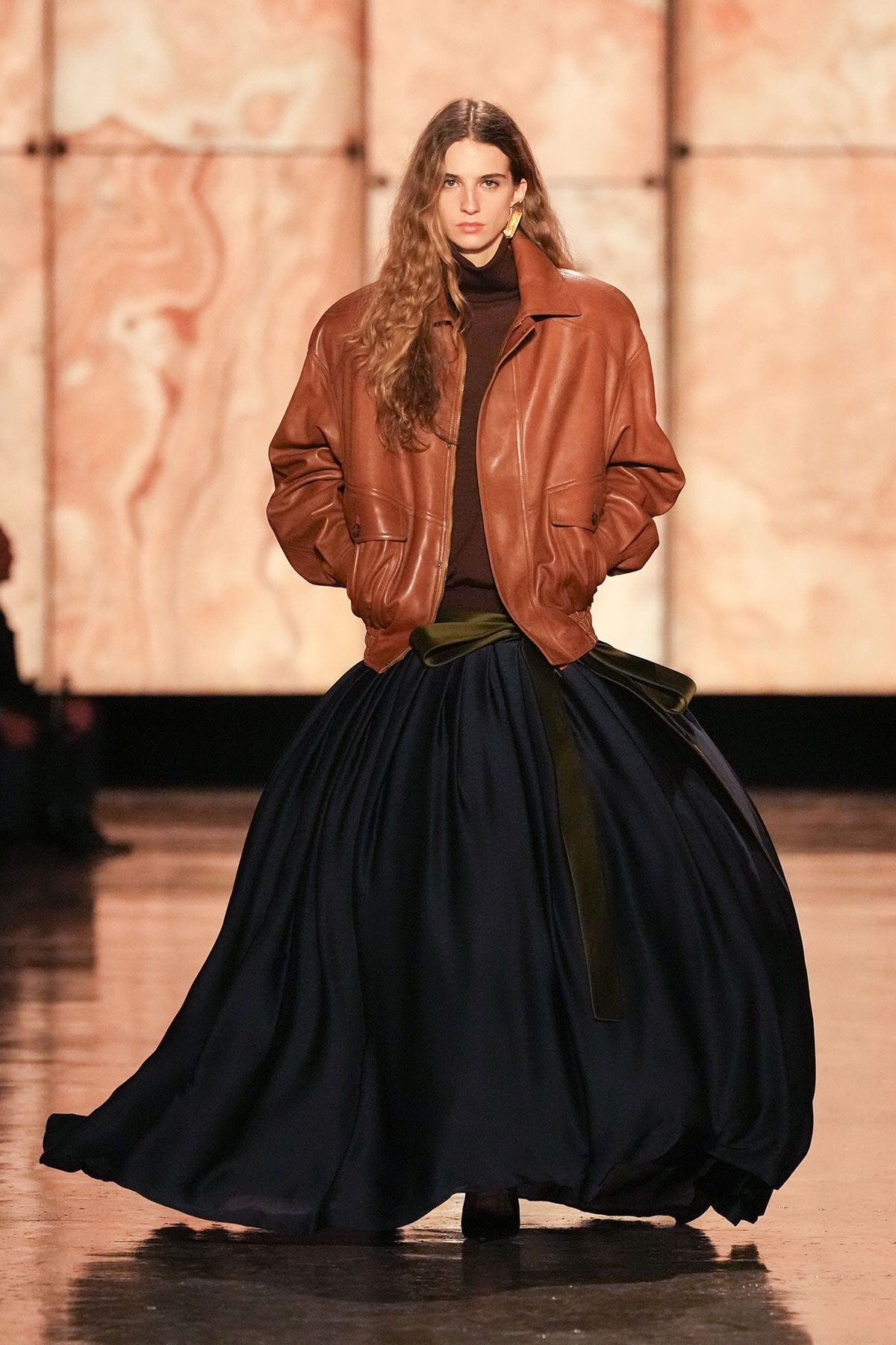 Model on a runway wearing a brown oversized leather jacket over a black turtleneck and a voluminous navy skirt.
