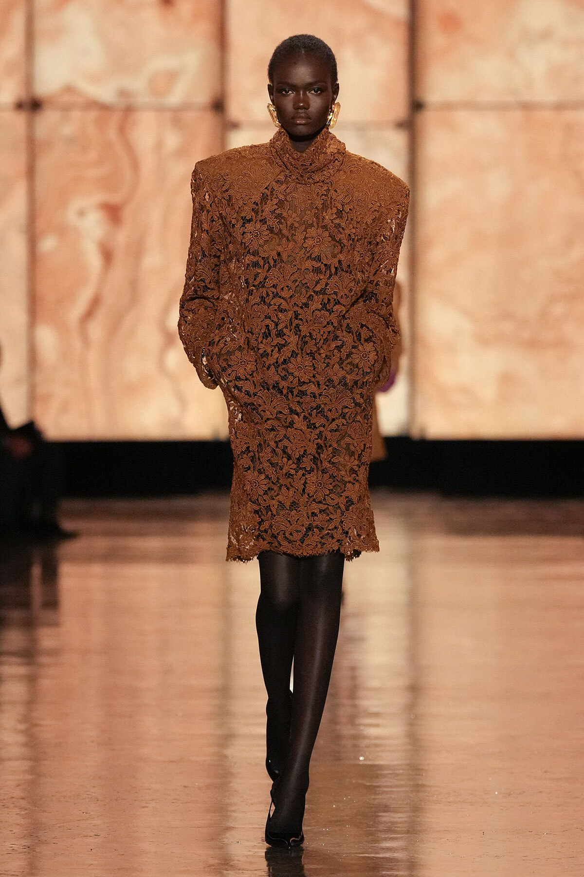 Model walking a runway in a brown floral lace knee-length dress with a high neckline, paired with black tights and heels, accessorized with large gold earrings.