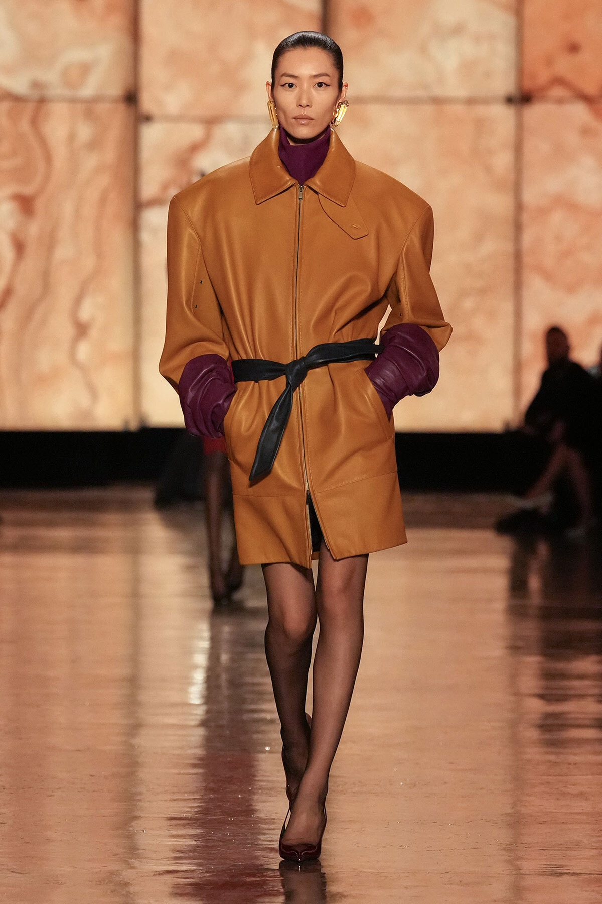 Model walks the runway in a camel leather coat with burgundy cuffs and a black belt at the waist.