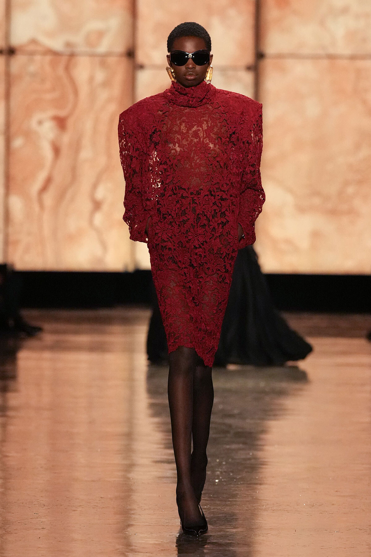 Model wearing a burgundy lace dress with a high neck and long sleeves, black sunglasses, and gold statement earrings on a runway.