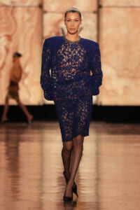Model walks runway in a navy blue lace knee-length dress with sheer floral pattern, black tights, and black heels.