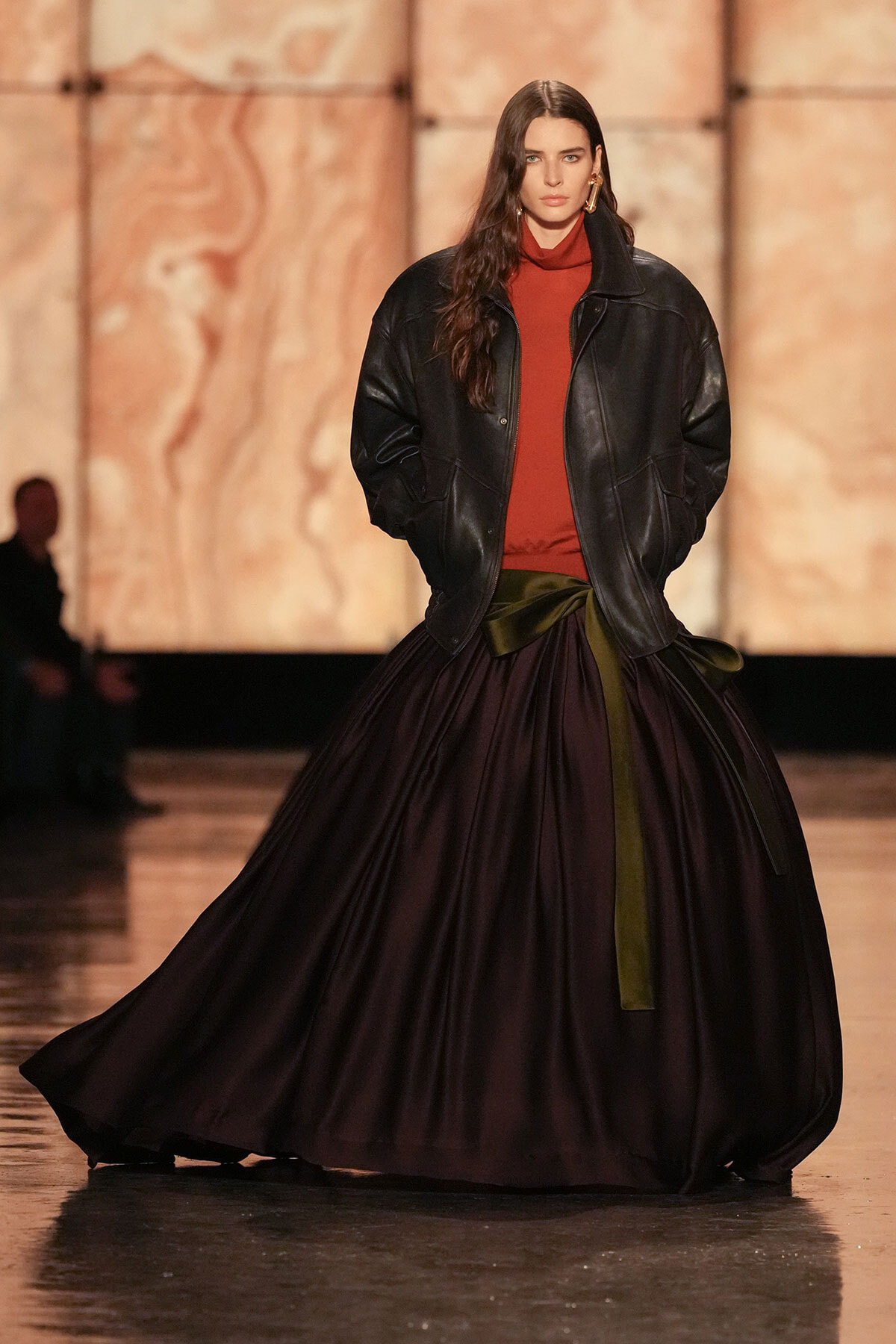 Runway model in an oversized black leather jacket over a rust turtleneck and a voluminous dark purple skirt with a green sash.