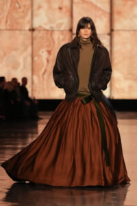 Model walking a fashion runway in an oversized black leather jacket, olive turtleneck, and a voluminous rust-colored skirt cinched with a dark belt.