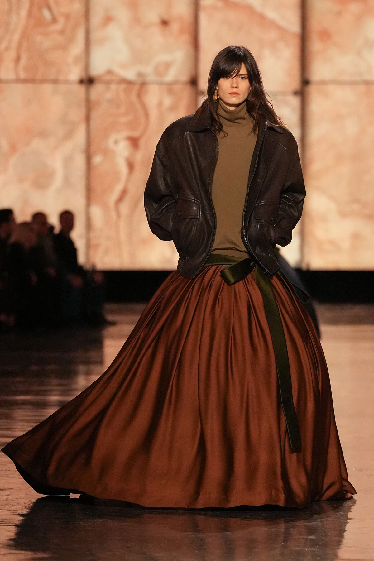 Model walking a fashion runway in an oversized black leather jacket, olive turtleneck, and a voluminous rust-colored skirt cinched with a dark belt.