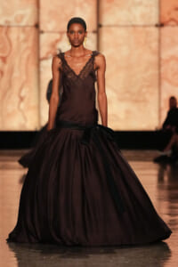 Model on a runway wearing a deep burgundy satin gown with a lace V-neck bodice and a large black sash bow at the waist.