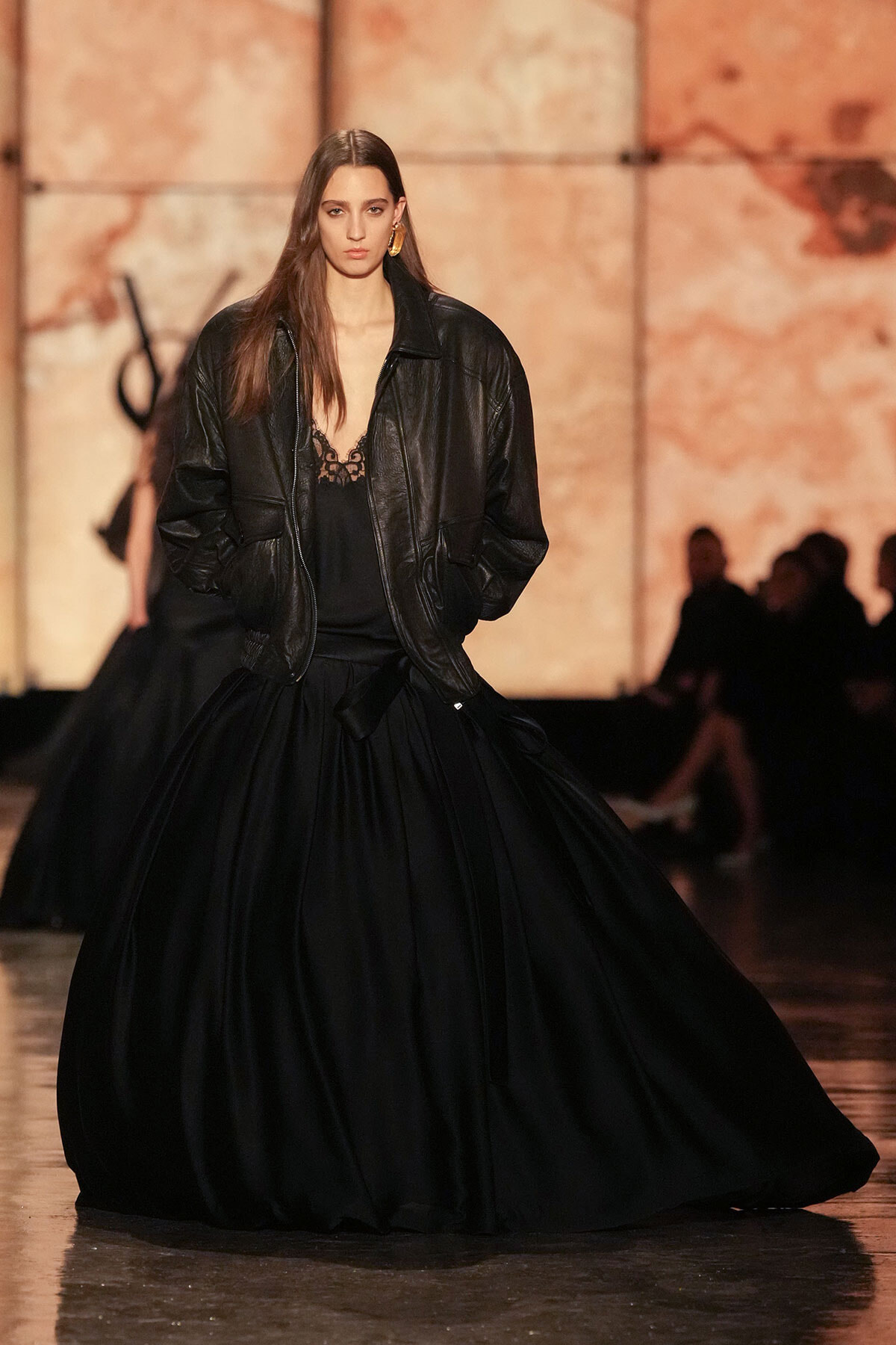 Model walking runway in oversized black leather jacket over a black lace-trim top and a voluminous black skirt.