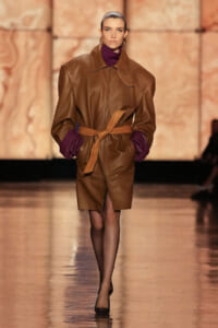 Model walks the runway in a oversized brown leather coat belted at the waist, with a purple turtleneck and purple sleeves showing at the cuffs.
