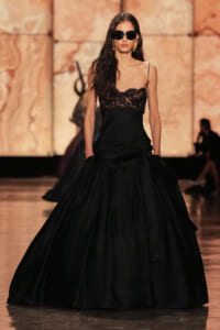 Model walks the runway in a black lace bodice gown with a full, voluminous skirt and dark sunglasses.