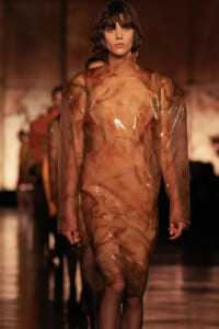 Model walking a runway in a sheer, amber-toned plastic dress with long sleeves and a knee-length hem.