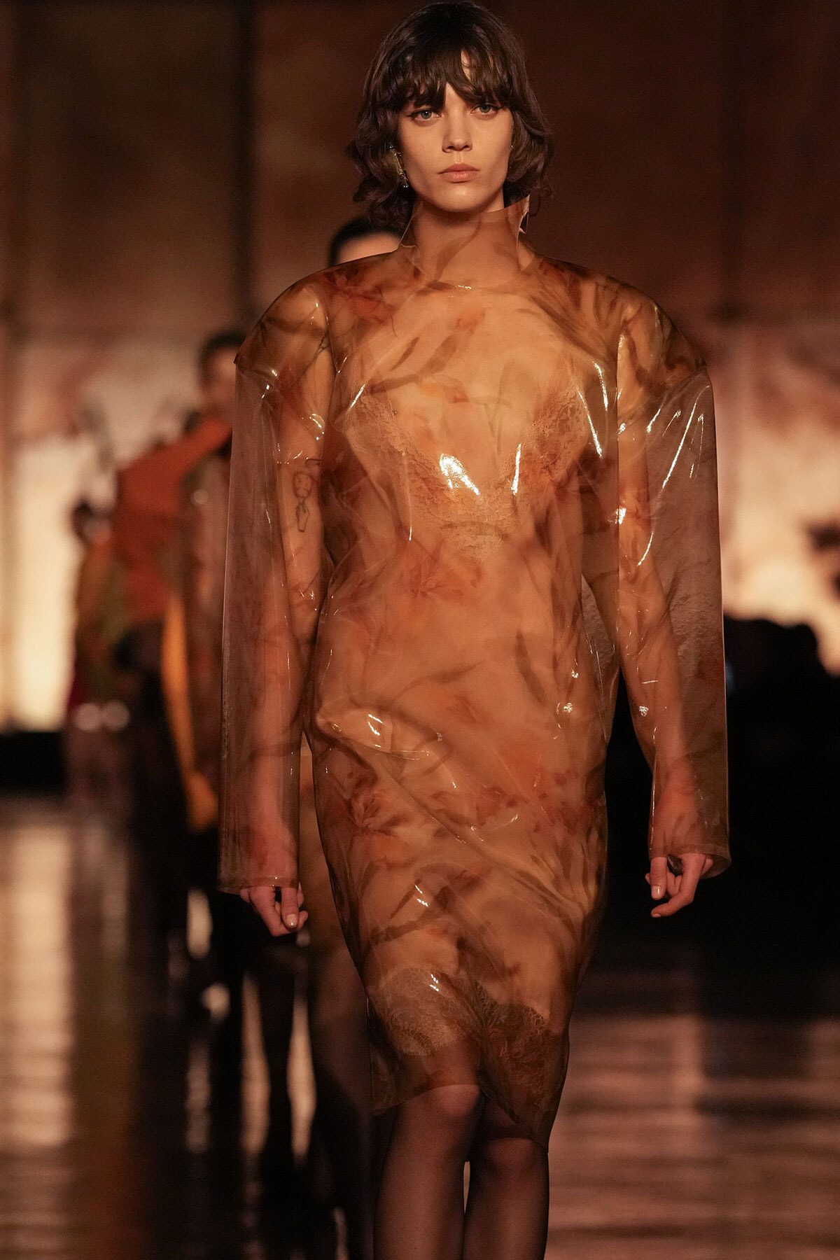 Model walking a runway in a sheer, amber-toned plastic dress with long sleeves and a knee-length hem.