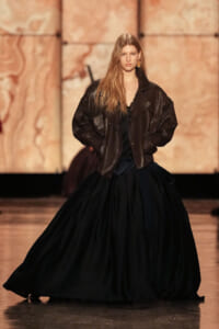 Model on a runway wearing an oversized black leather jacket over a full, voluminous black dress, hands in pockets.