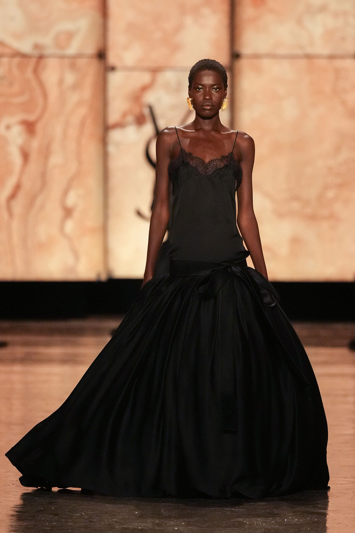 Tall model walking the runway in a black lace-trim evening gown with a voluminous skirt and gold statement earrings.