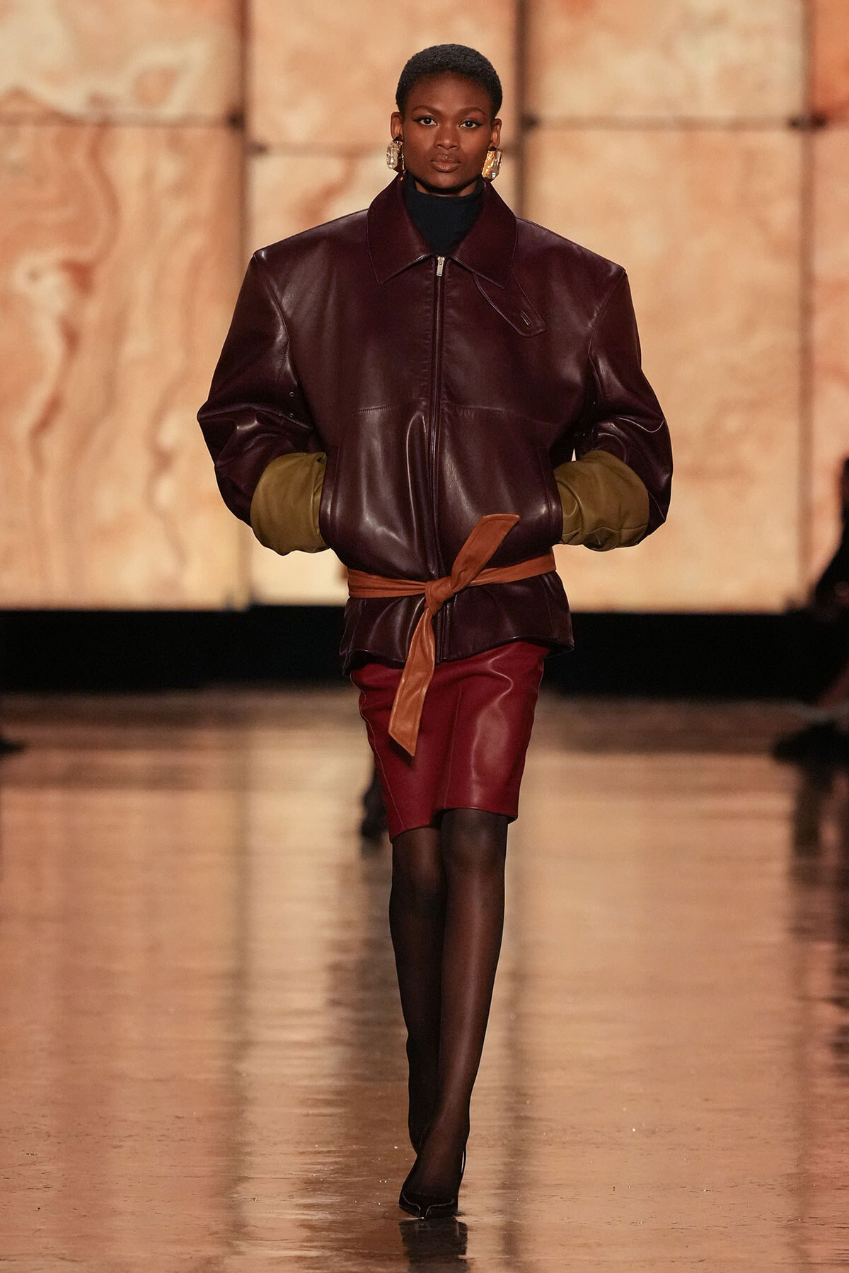 Model on a runway wearing an oversized burgundy leather jacket with olive-green cuffs, a tan belt, and a matching burgundy pencil skirt, paired with black tights and heels.