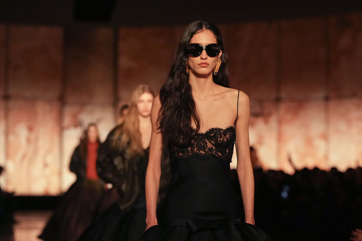 Model walking a fashion runway in a black lace-detailed gown and oversized sunglasses, spotlight on her as audience watches in the background.