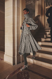 Person wearing a long grey polka-dot trench coat with a high collar, stepping down marble stairs in a grand interior.