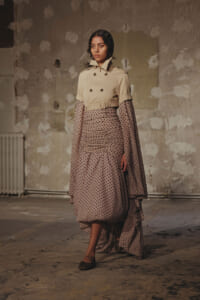 Female model wearing a beige double‑breasted trench over a polka-dot, ruched,-layered midi dress, posing in a distressed studio backdrop.