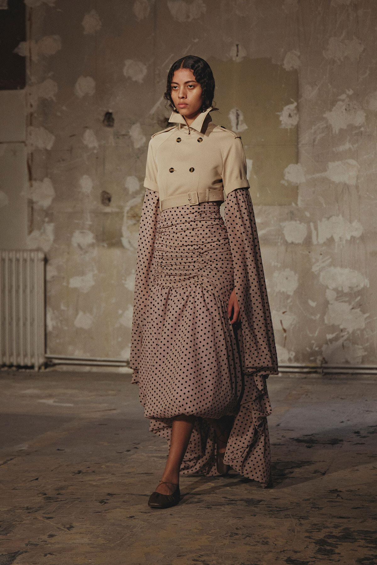 Female model wearing a beige double‑breasted trench over a polka-dot, ruched,-layered midi dress, posing in a distressed studio backdrop.