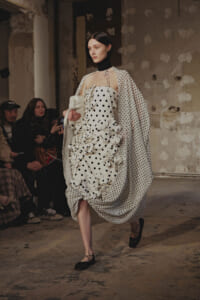 Model walks the runway in a white polka-dot dress with sculpted ruffles and a matching dotted cape in an industrial venue.