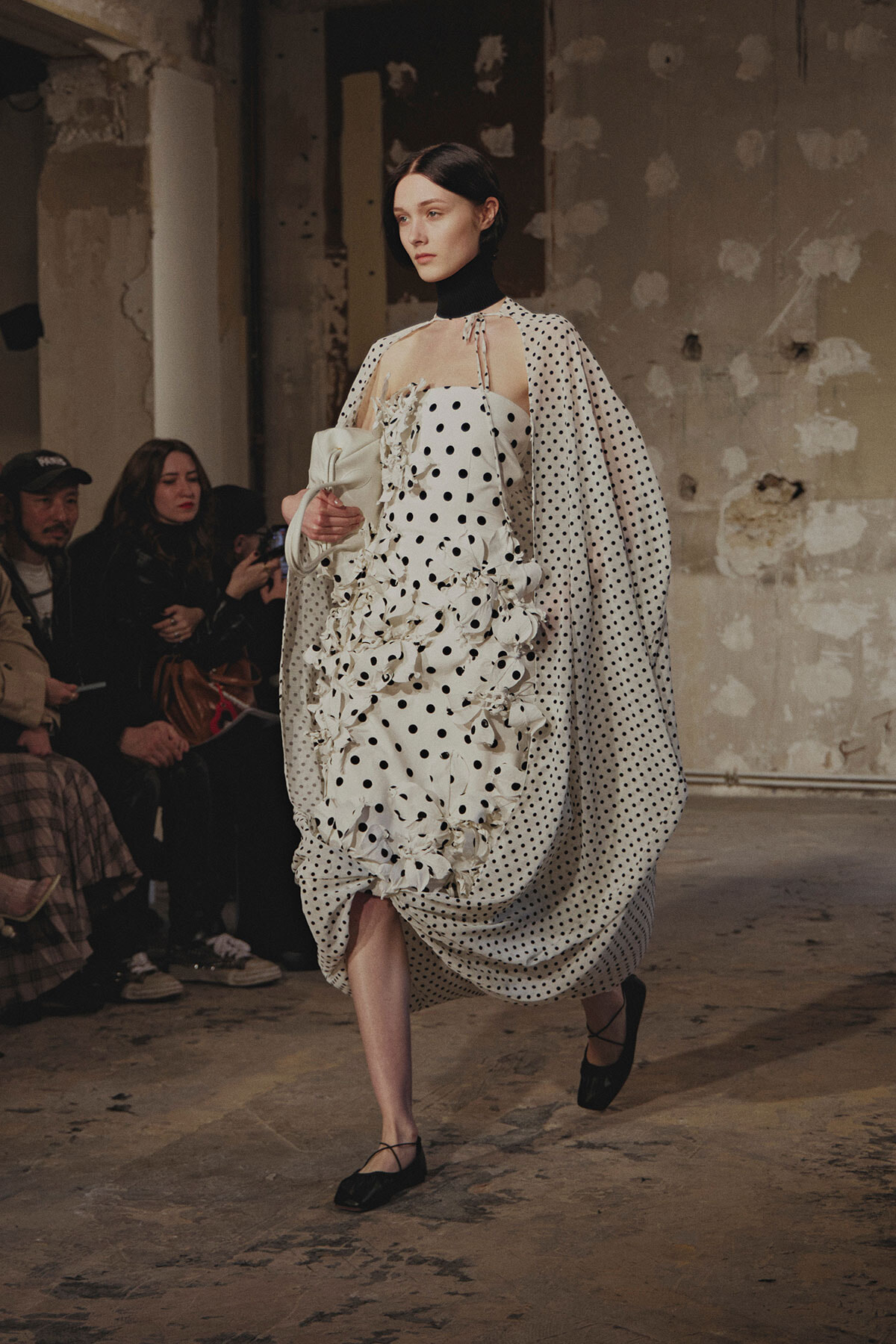 Model walks the runway in a white polka-dot dress with sculpted ruffles and a matching dotted cape in an industrial venue.