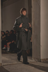 Male model walking a runway in a double-breasted gray wool coat with a belt and high collar.
