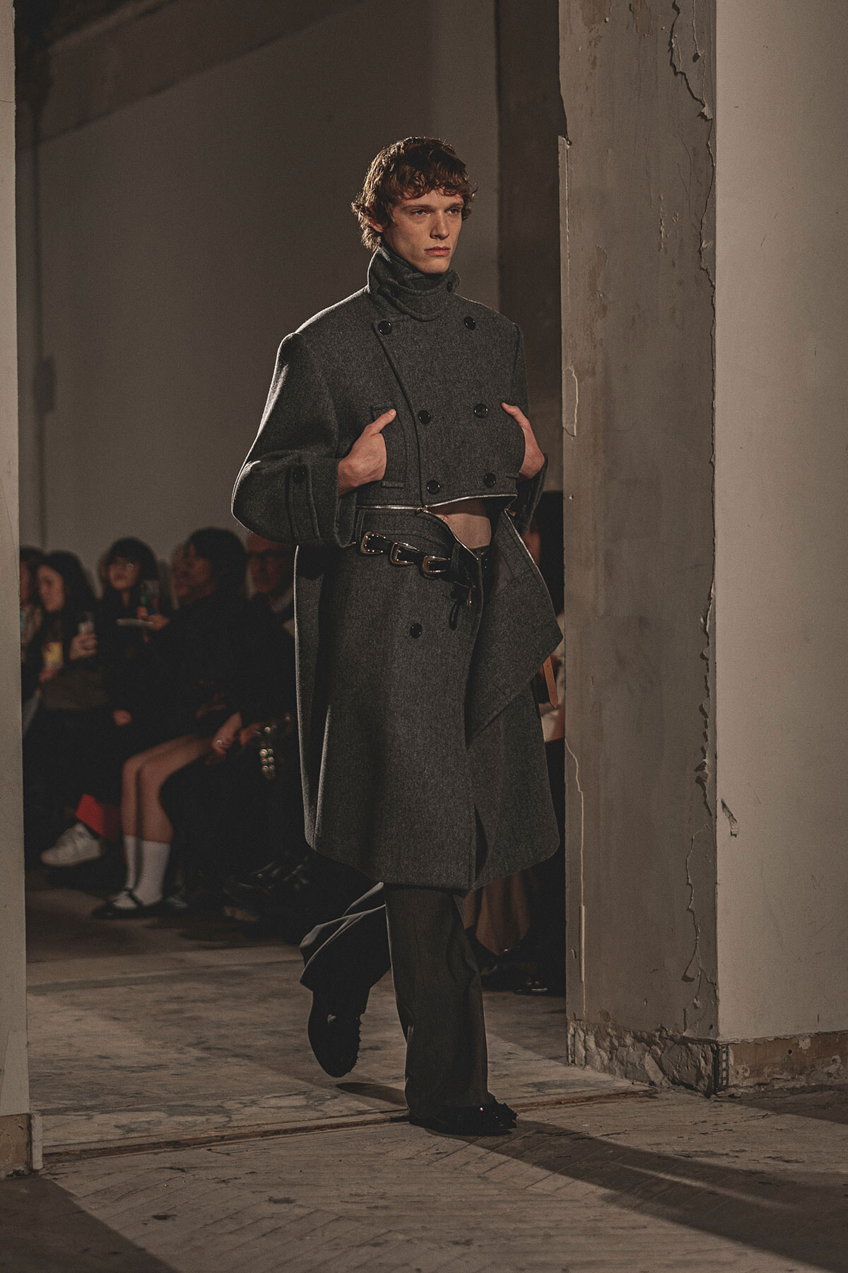 Male model walking a runway in a double-breasted gray wool coat with a belt and high collar.