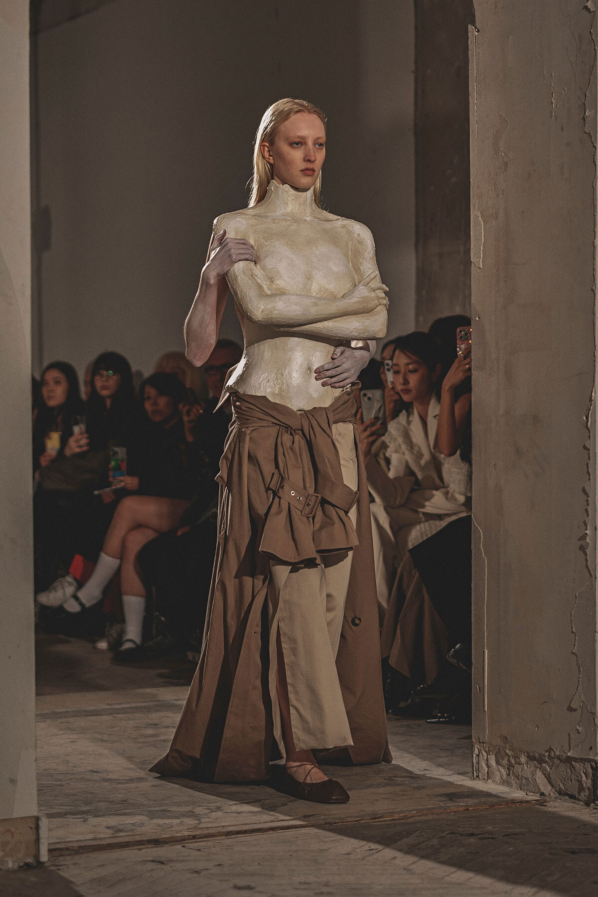 Model on a runway wearing a white sculpted turtleneck top and a layered tan skirt with a tied belt, audience in the background.