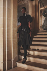Black woman in a black haute-couture dress with dramatic puff sleeves, standing on a stone staircase in a historic building.