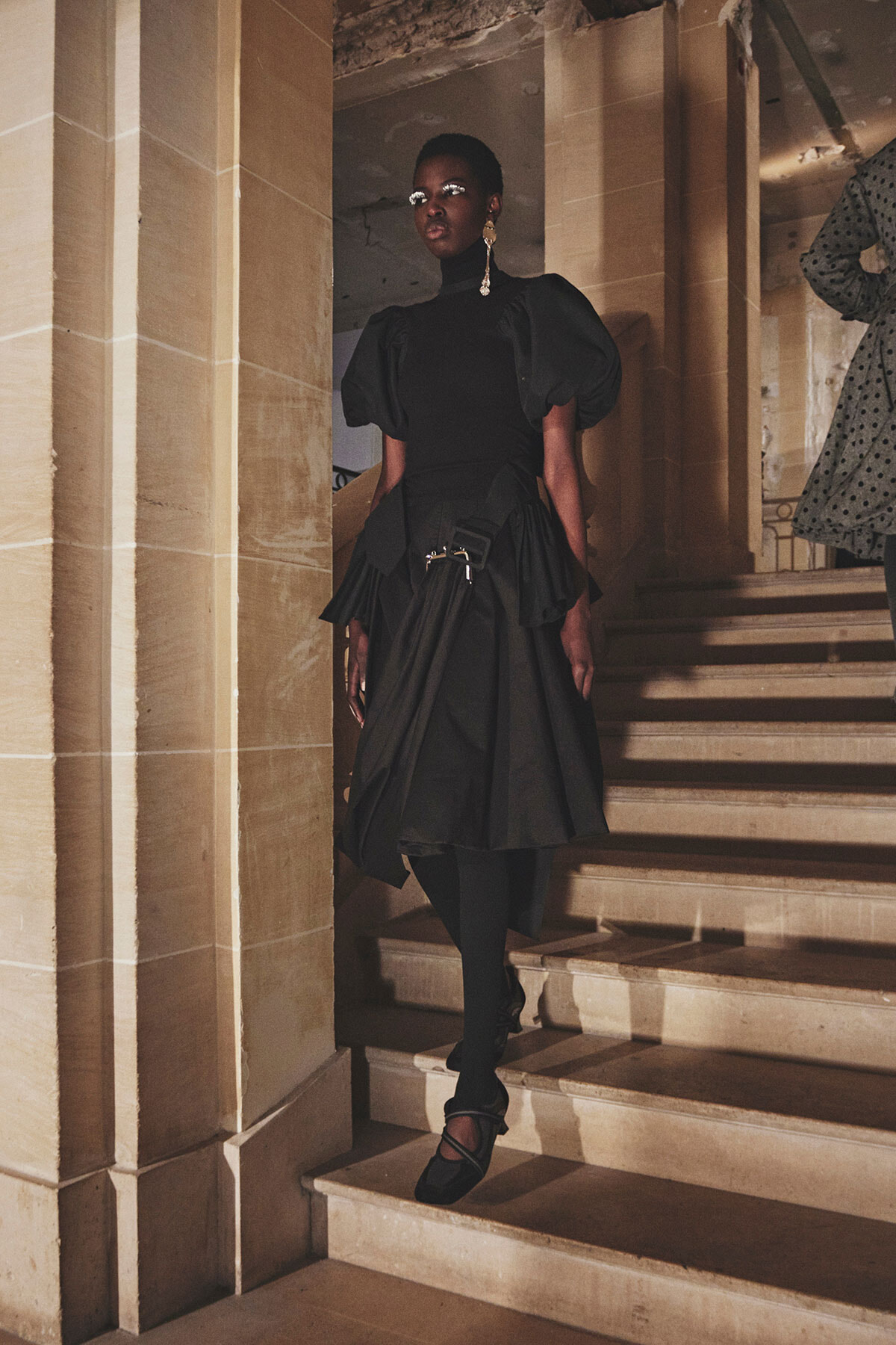 Black woman in a black haute-couture dress with dramatic puff sleeves, standing on a stone staircase in a historic building.