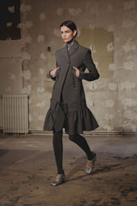 Model in a gray, double-breasted coatdress with rows of buttons and a ruffled hem, standing in a worn industrial studio.