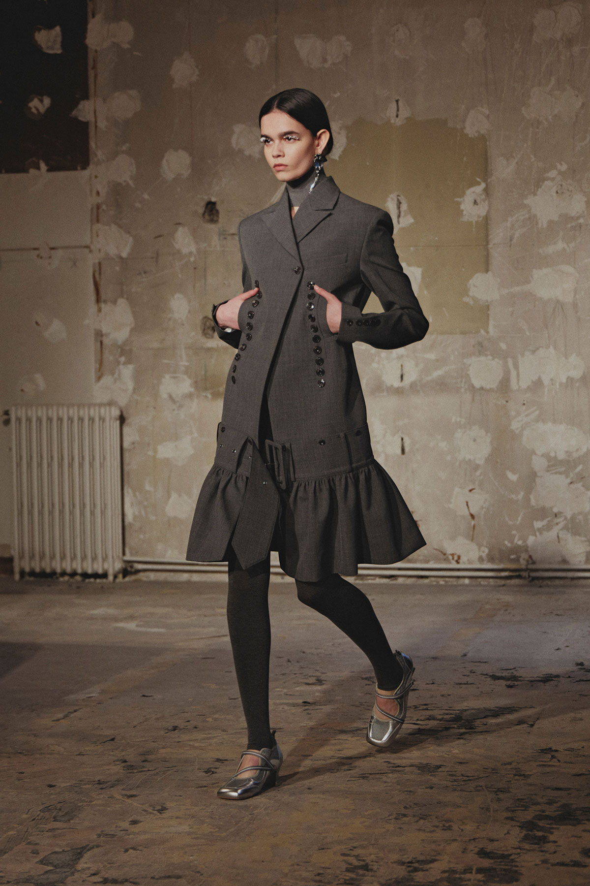 Model in a gray, double-breasted coatdress with rows of buttons and a ruffled hem, standing in a worn industrial studio.