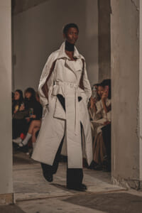 Model walks a runway in an oversized white trench coat with a belt, black turtleneck, and cape-like sleeves, audience watching in the background.
