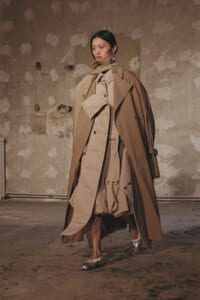 Model walks in a layered tan trench coat ensemble in a bare, textured studio with peeling walls, wearing metallic shoes and statement earrings