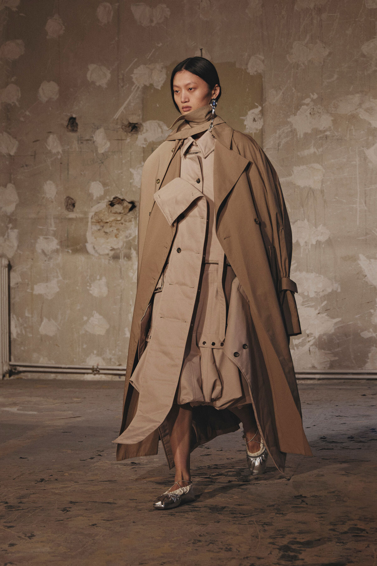 Model walks in a layered tan trench coat ensemble in a bare, textured studio with peeling walls, wearing metallic shoes and statement earrings