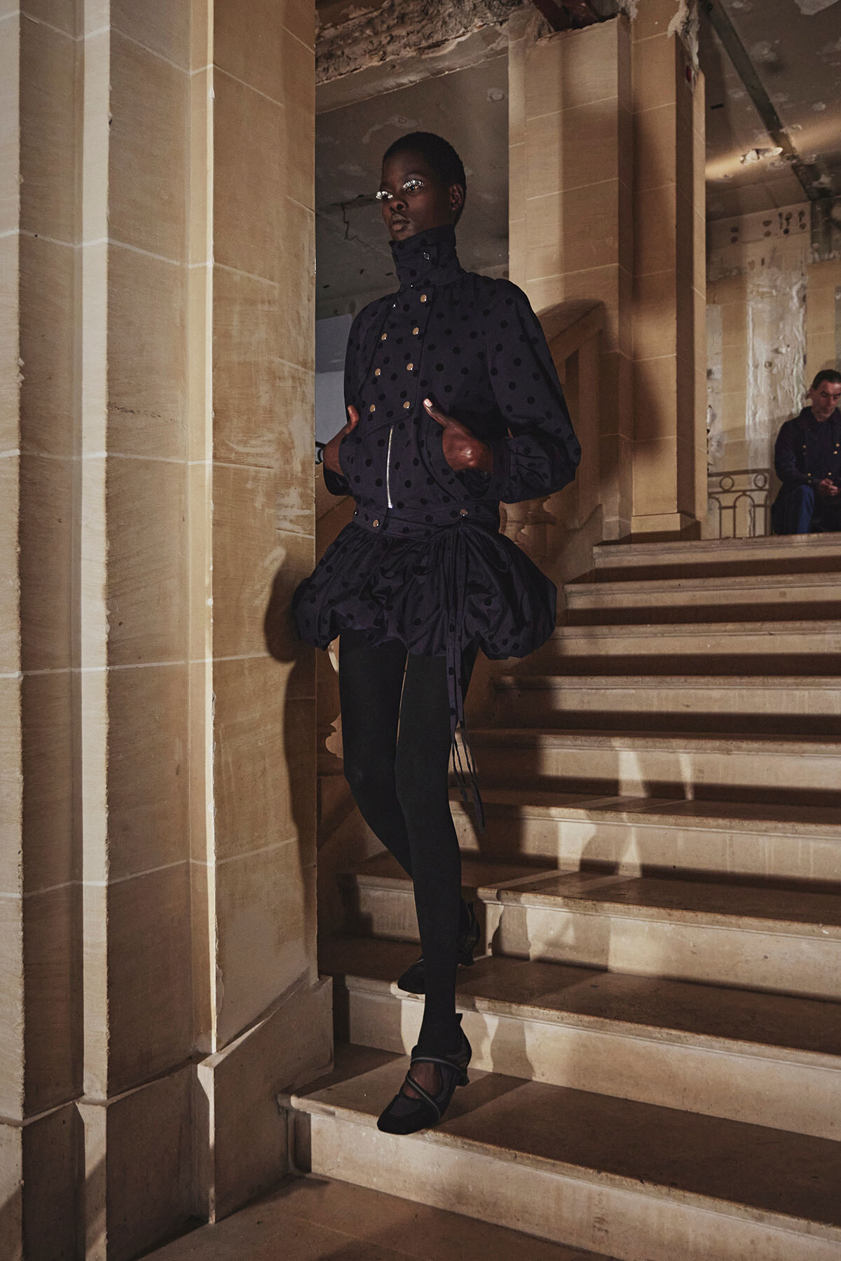 Model wearing a dark navy polka-dot jacket with a voluminous bubble skirt, descending a marble staircase in a dim historic interior.